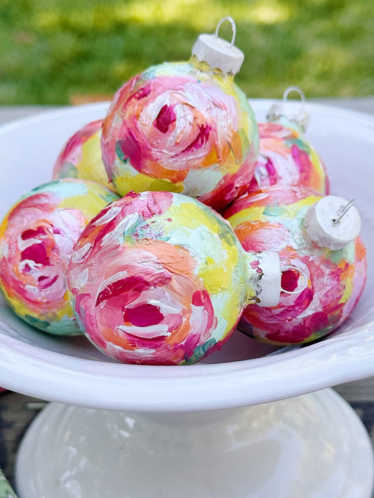 A white bowl holds several round ornaments decorated with bright, multicolored abstract floral patterns. Grass is visible in the background.