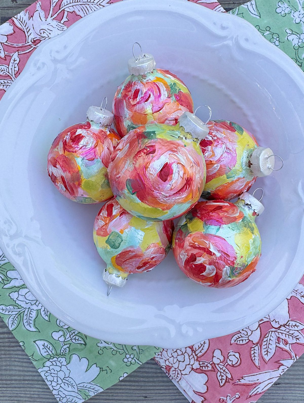 A white bowl holds six colorful, hand-painted Christmas ornaments with floral patterns, placed on patterned pink and green napkins.
