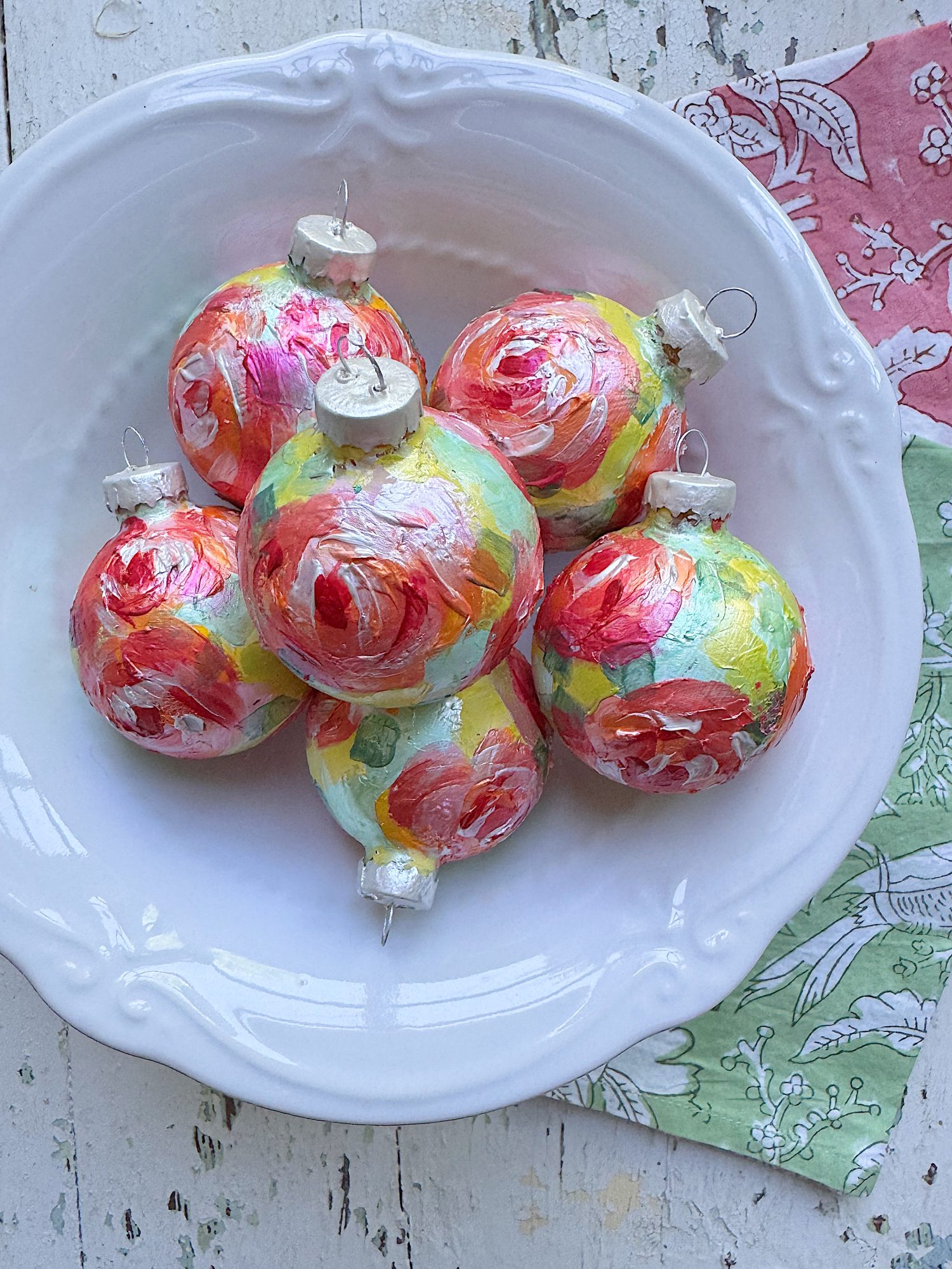 Six colorful hand-painted glass ornaments in red, pink, yellow, and green shades arranged in a white decorative bowl on a rustic surface.
