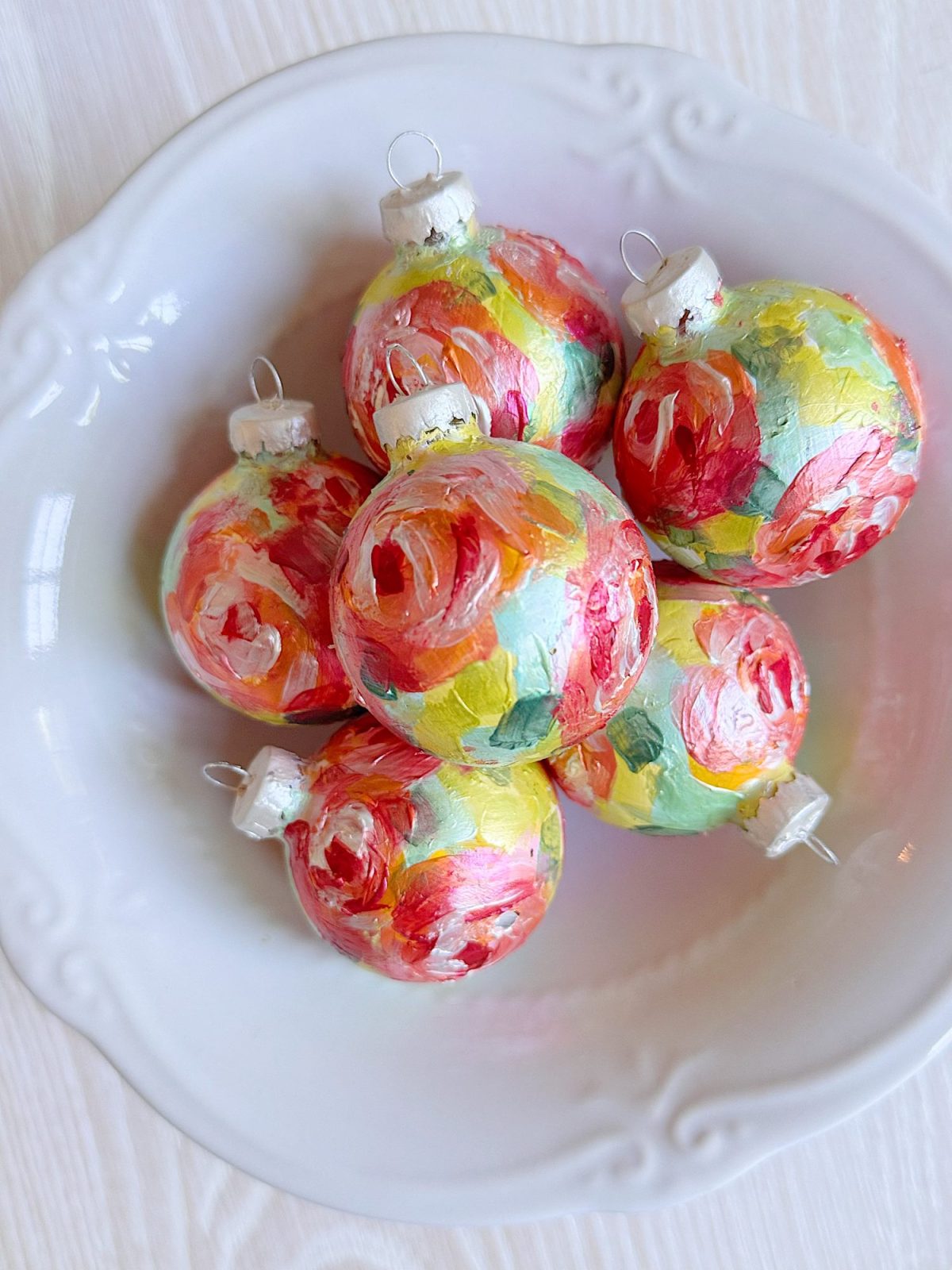 A white bowl containing six Christmas ornaments painted with bright floral designs in red, pink, yellow, and green.