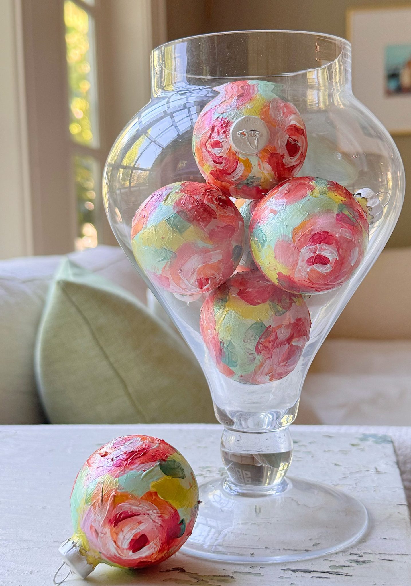A glass vase holds five hand-painted, pastel swirl ornaments, with one similar ornament resting on the table beside it.