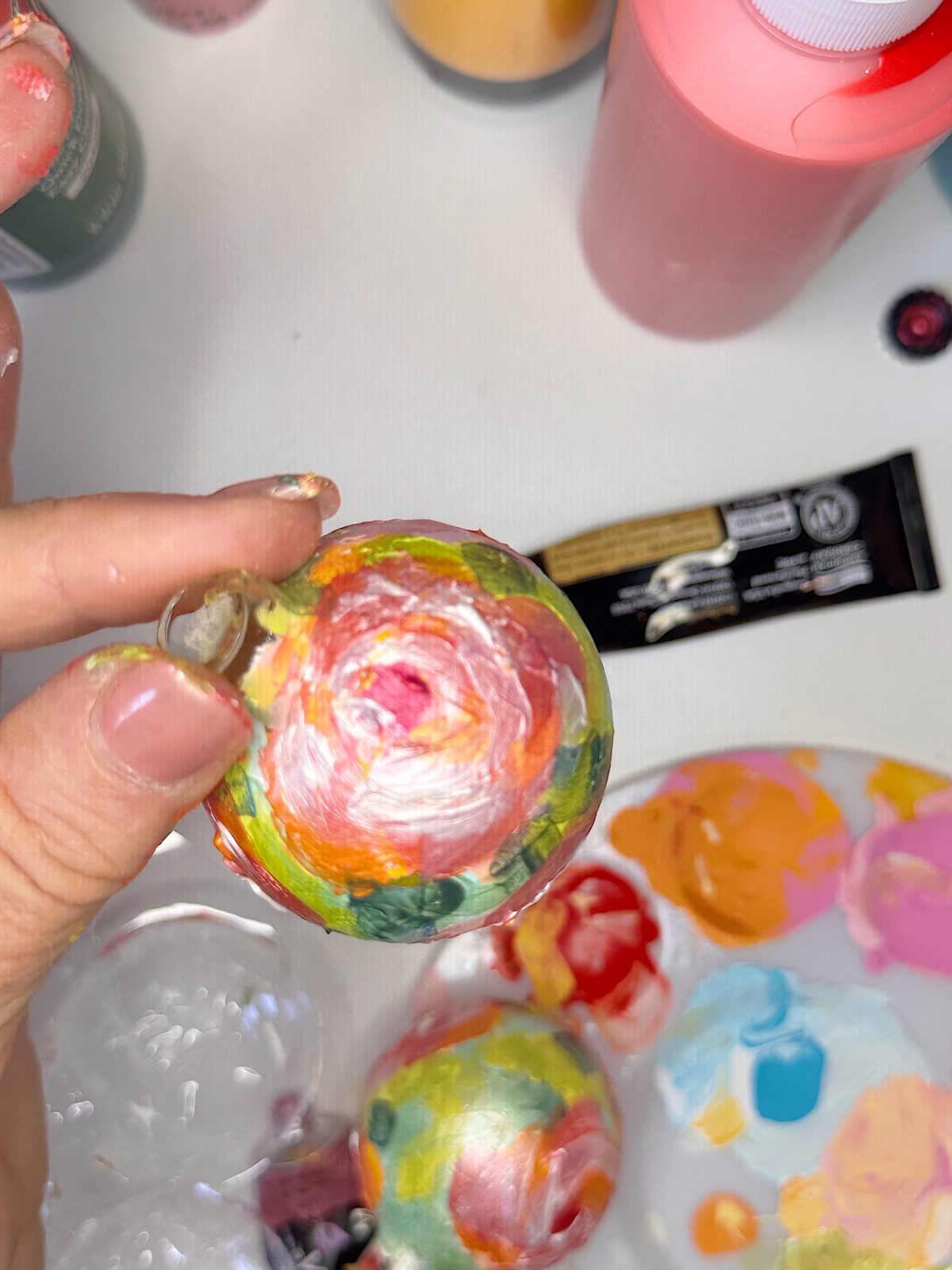 A hand holds a round ornament painted with colorful floral patterns; various paints and a palette with mixed colors are visible on the table.