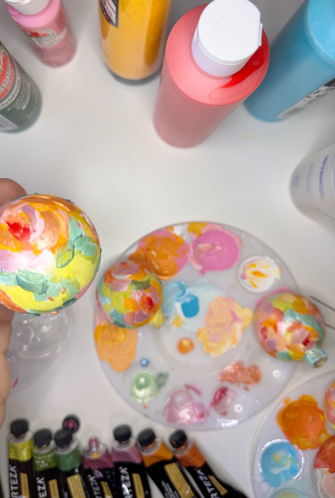 Hand holding a painted ornament above a palette with colorful paint blobs, surrounded by paint bottles and tubes on a white surface.