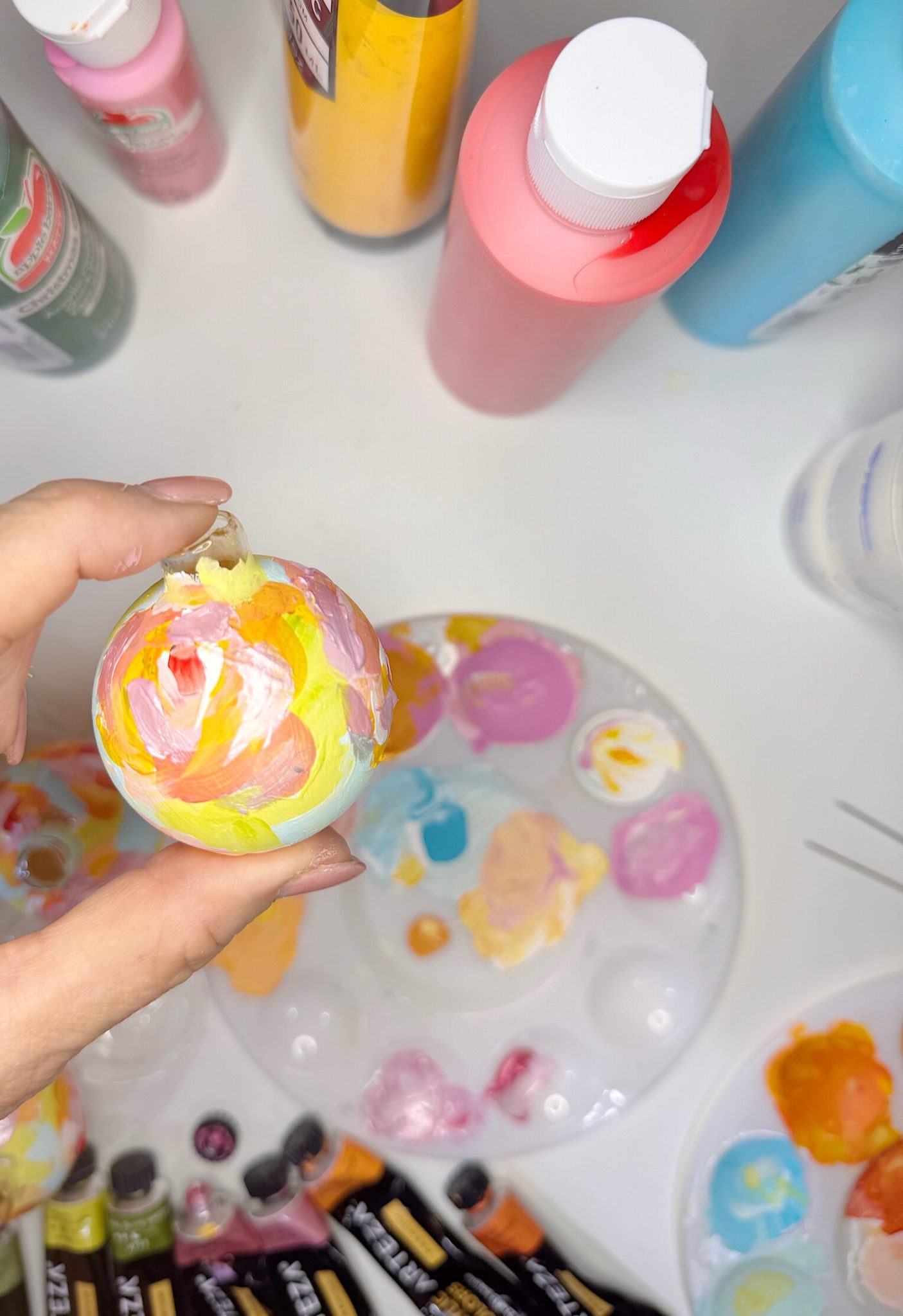 A hand holds a small round object painted with pink, yellow, and white, above a palette with mixed paints and various bottles and tubes of paint on a white surface.