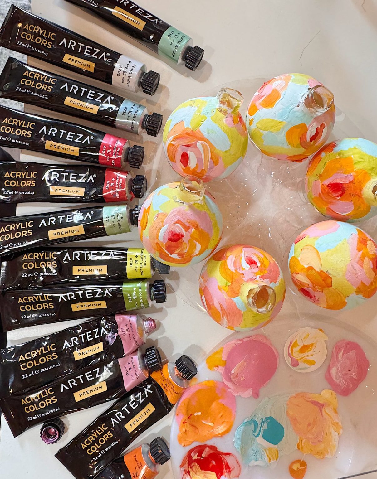 Several tubes of Arteza acrylic paint are arranged beside round glass ornaments painted with colorful abstract patterns in pink, yellow, orange, and white tones. Paint blobs are visible nearby.
