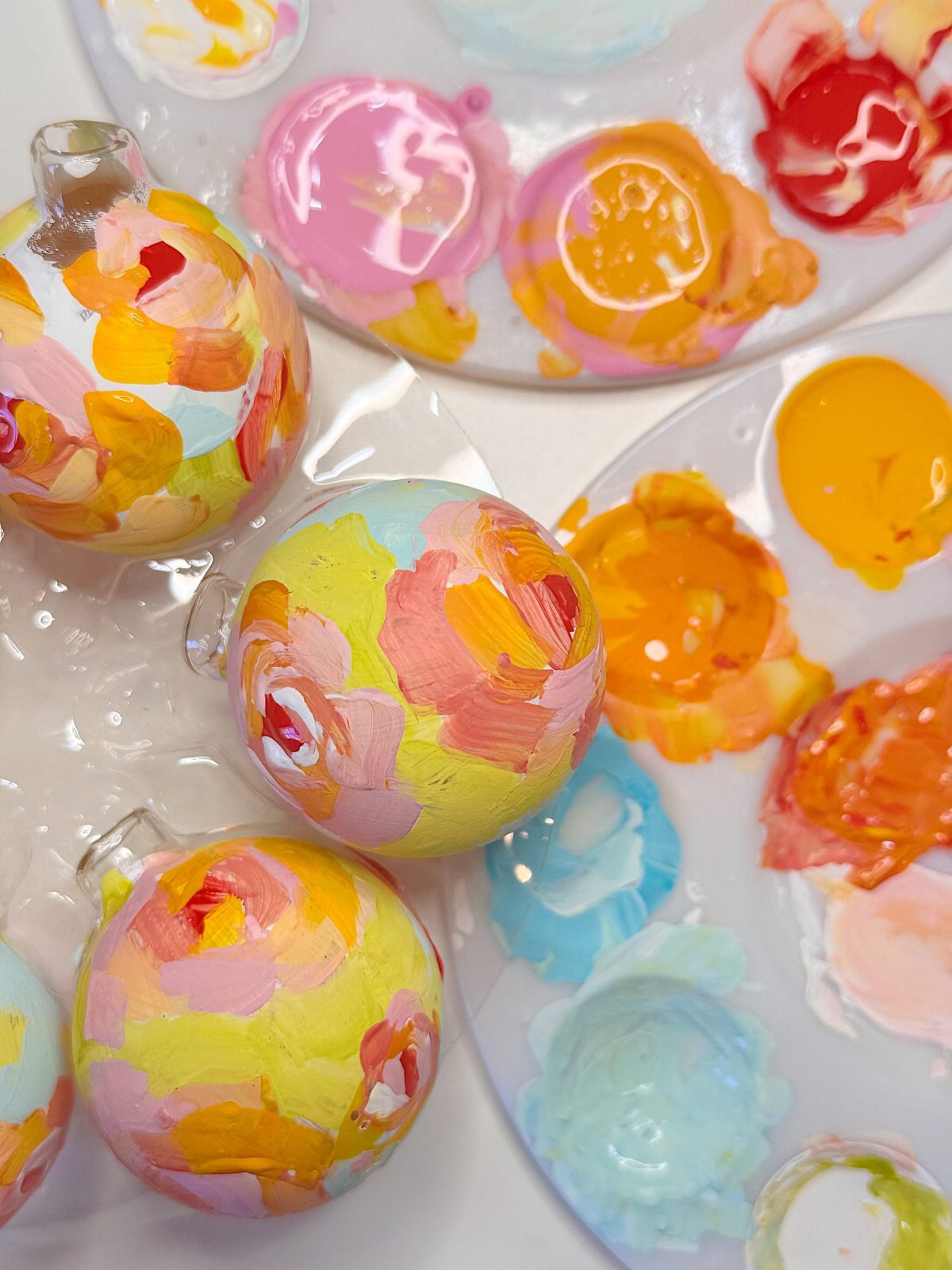 Three glass ornaments painted with colorful, abstract brushstrokes rest beside palettes holding various pastel and bright paint blobs.