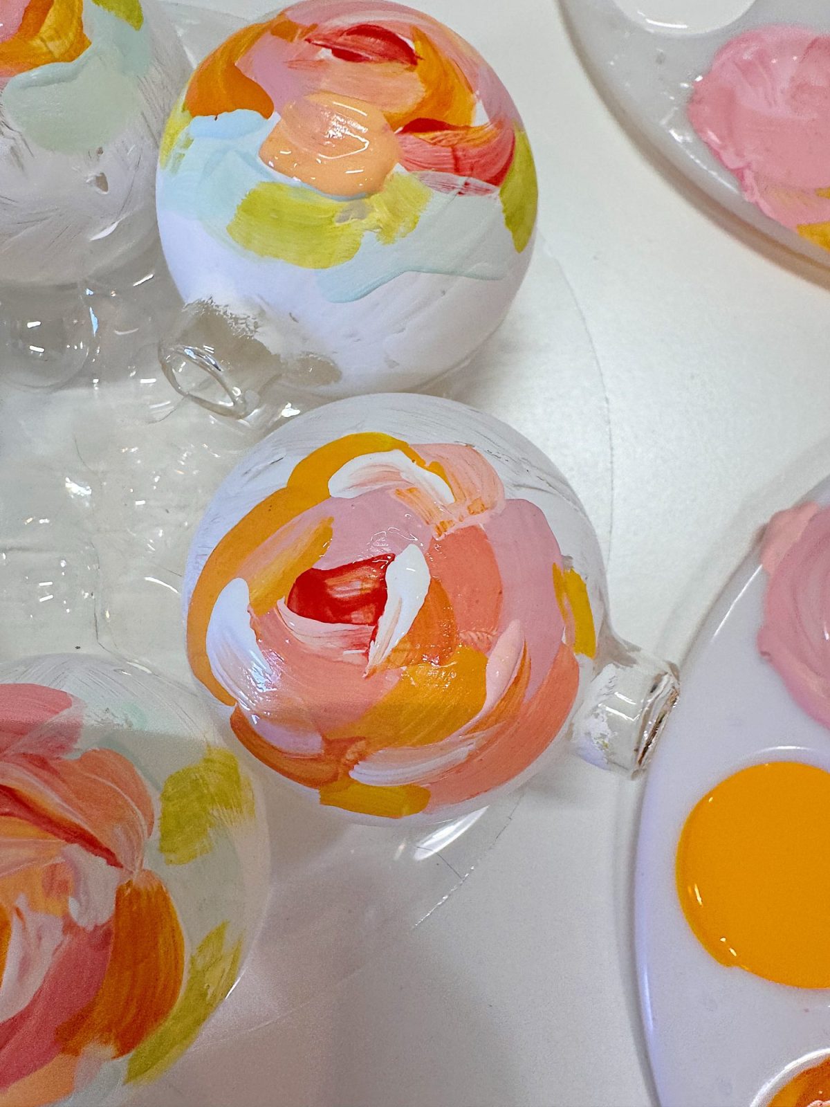 A white glass ornament is painted with pink, orange, yellow, and green brushstrokes, surrounded by a paint palette and other painted ornaments.