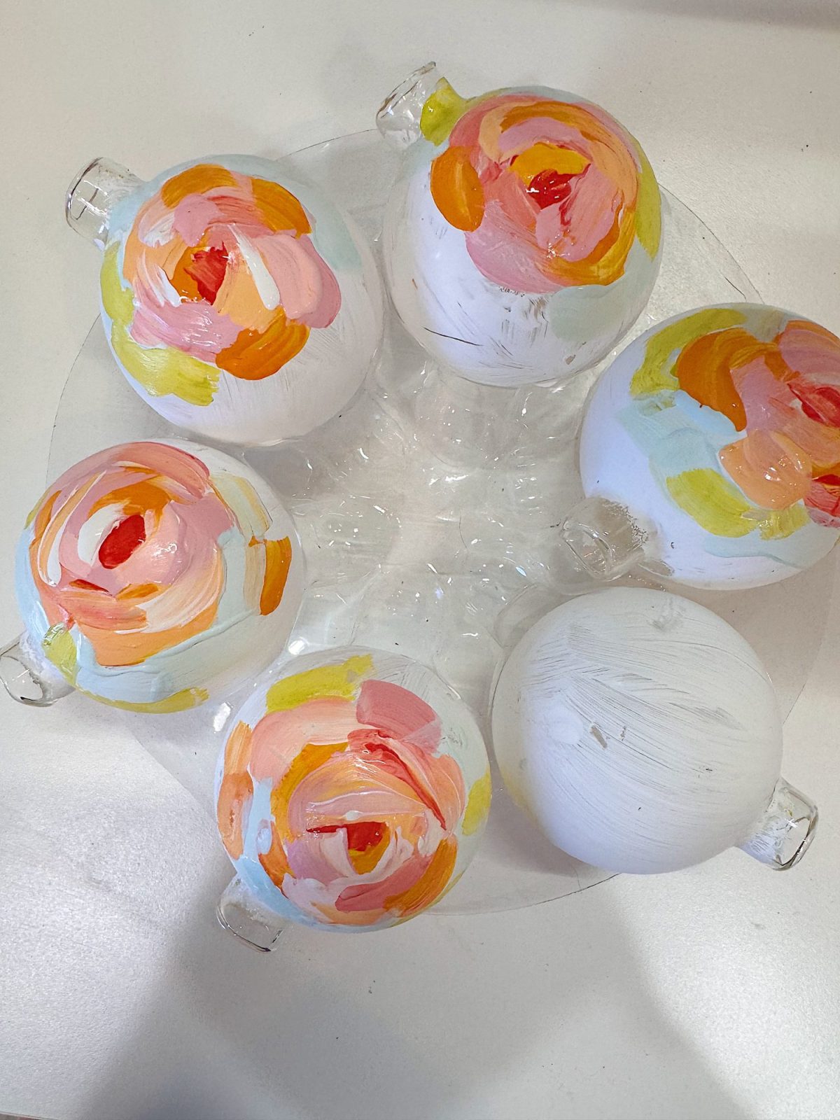 Six white glass ornaments with hand-painted floral designs in pink, orange, yellow, and light blue are arranged in a clear plastic holder on a white surface.