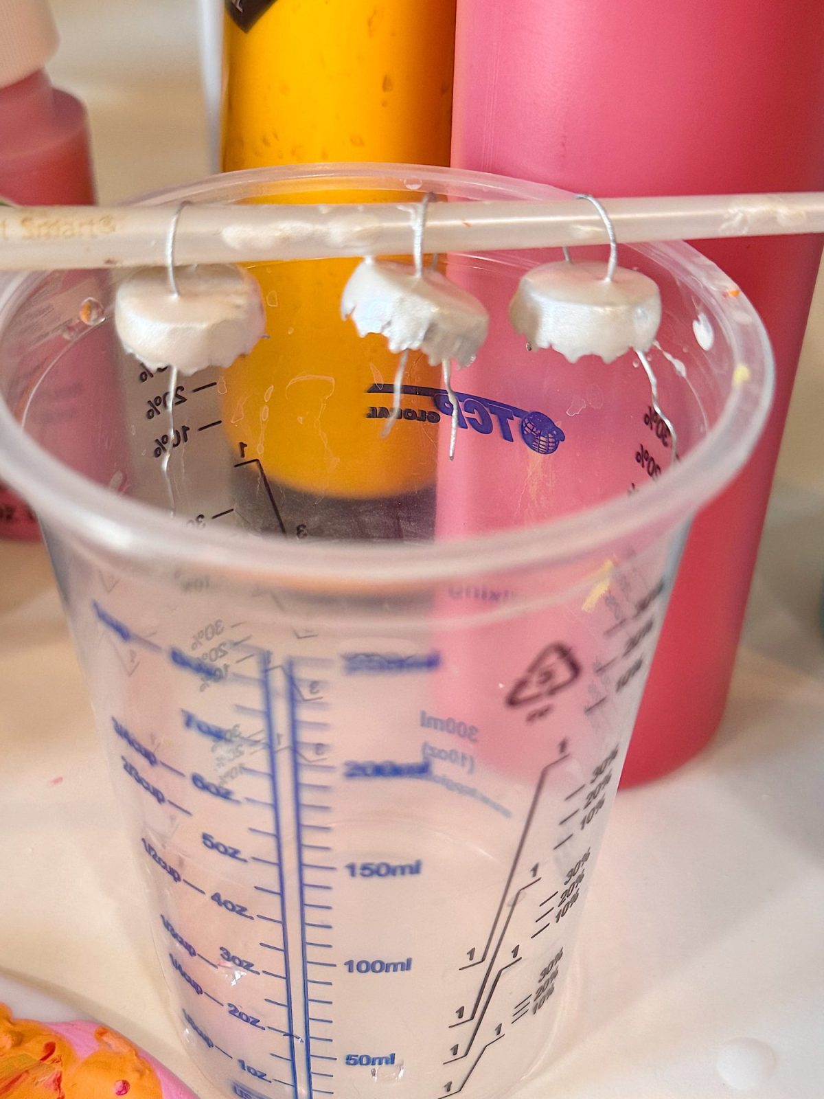 A clear plastic measuring cup with clay rings hanging on a straw above it, surrounded by bottles of yellow and pink paint.