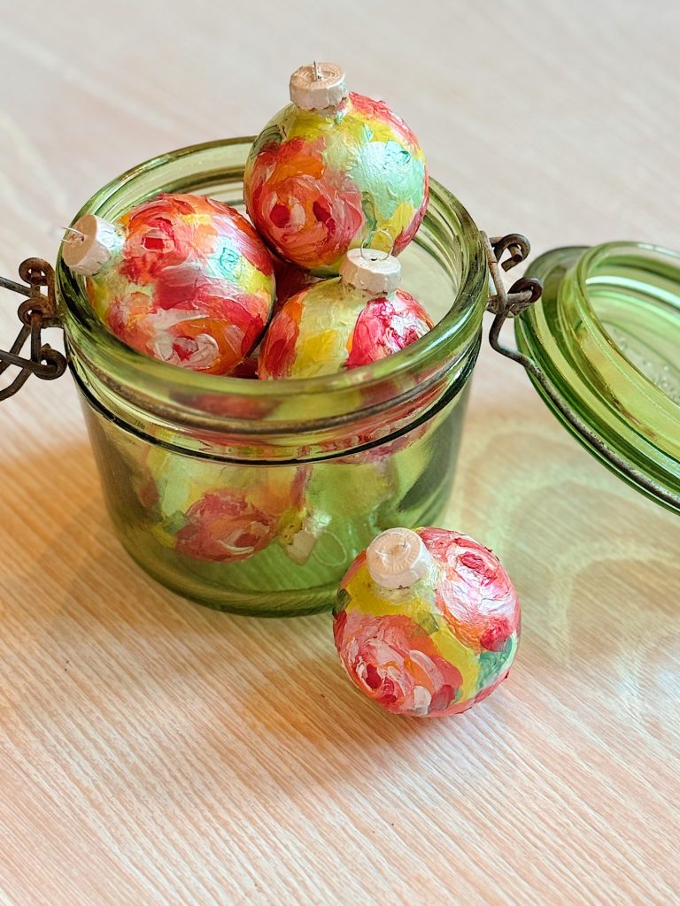 A green glass jar filled with colorful, hand-painted round ornaments, with one ornament resting outside the jar on a light wooden surface.