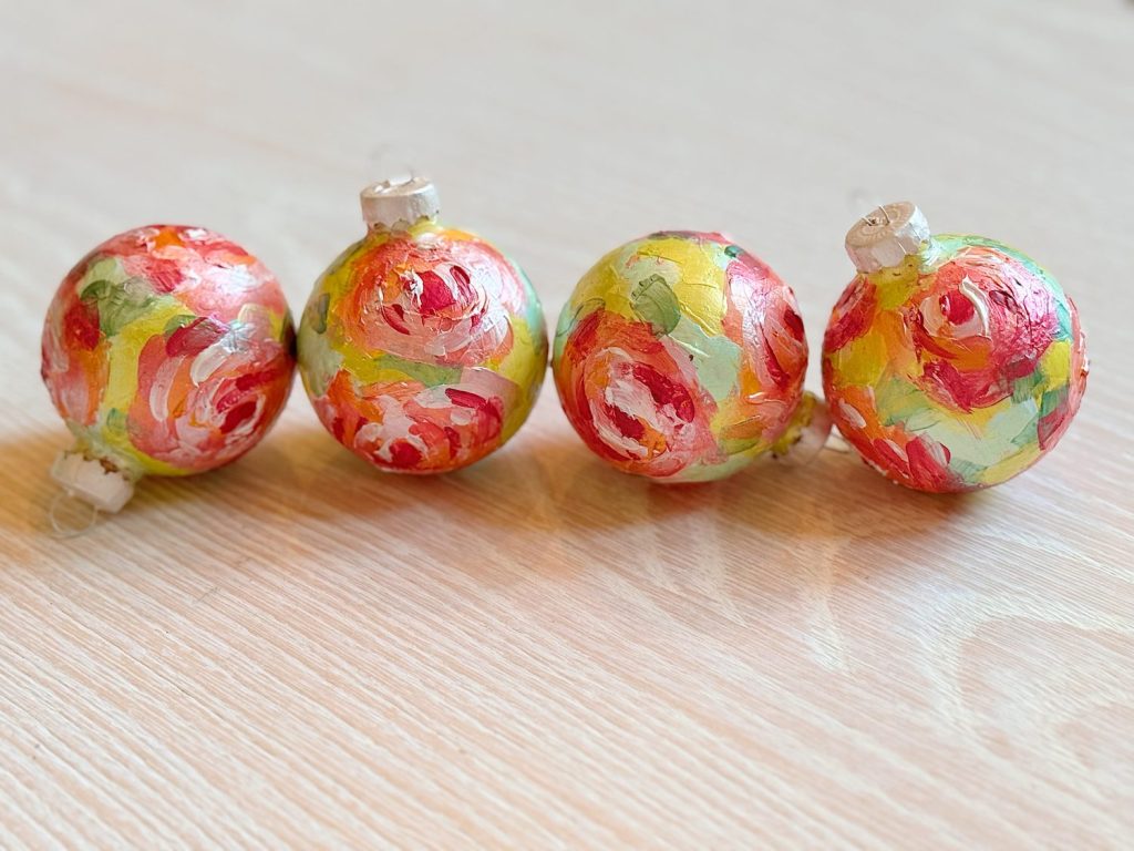 Four hand-painted Christmas ornaments with red, green, and yellow floral patterns arranged in a row on a light wooden surface.