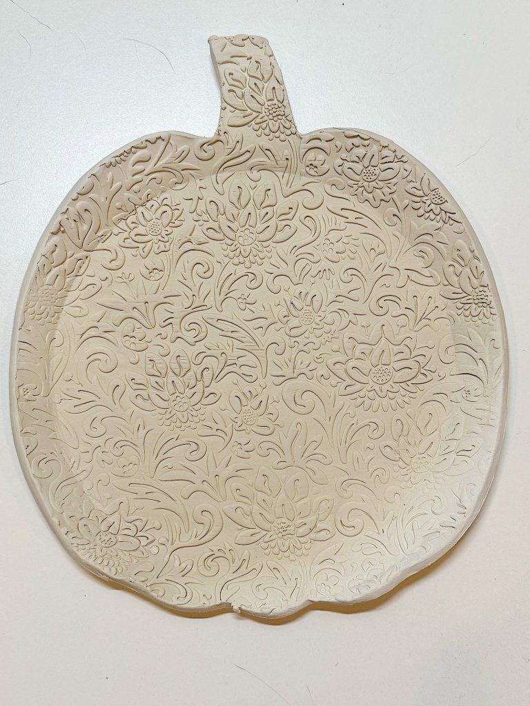 A ceramic plate shaped like a pumpkin with an intricate floral and leaf pattern embossed on its surface, placed on a white background.
