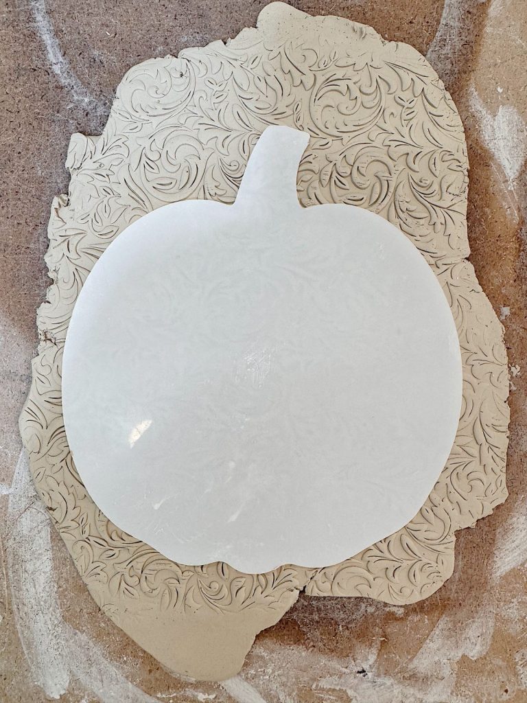 A sheet of clay with a floral pattern is cut into the shape of a pumpkin, placed on a textured surface.