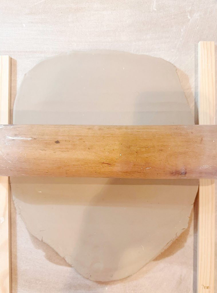 A rolling pin flattens a circular piece of clay between two wooden guides on a light-colored surface.