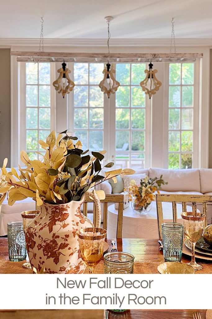 A family room decorated for fall with gold and brown accents, a floral vase with autumn foliage on the table, and large windows in the background letting in natural light.