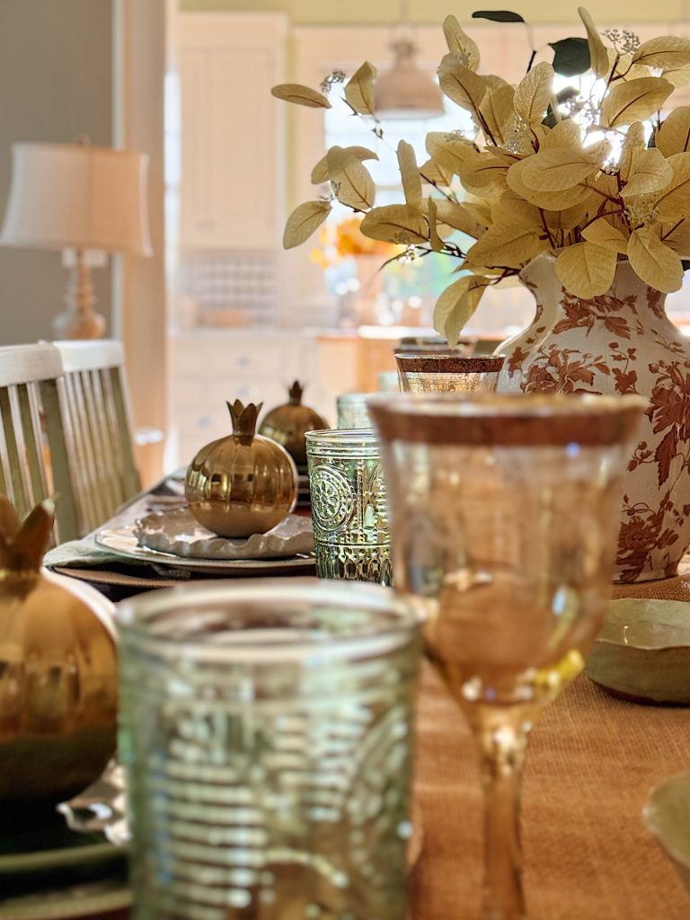 A dining table set with glassware, gold pomegranate decorations, and a large vase with beige foliage in a bright, sunlit room.