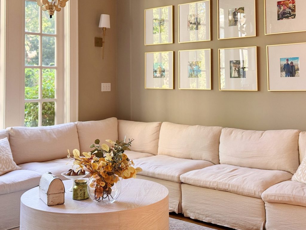 A living room with a white sectional sofa, round white coffee table with a floral arrangement, and framed photos on a beige wall near large windows.