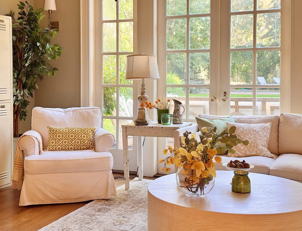 A bright living room with a white armchair, a beige sofa, a round wooden coffee table with yellow flowers, and large windows letting in natural light.