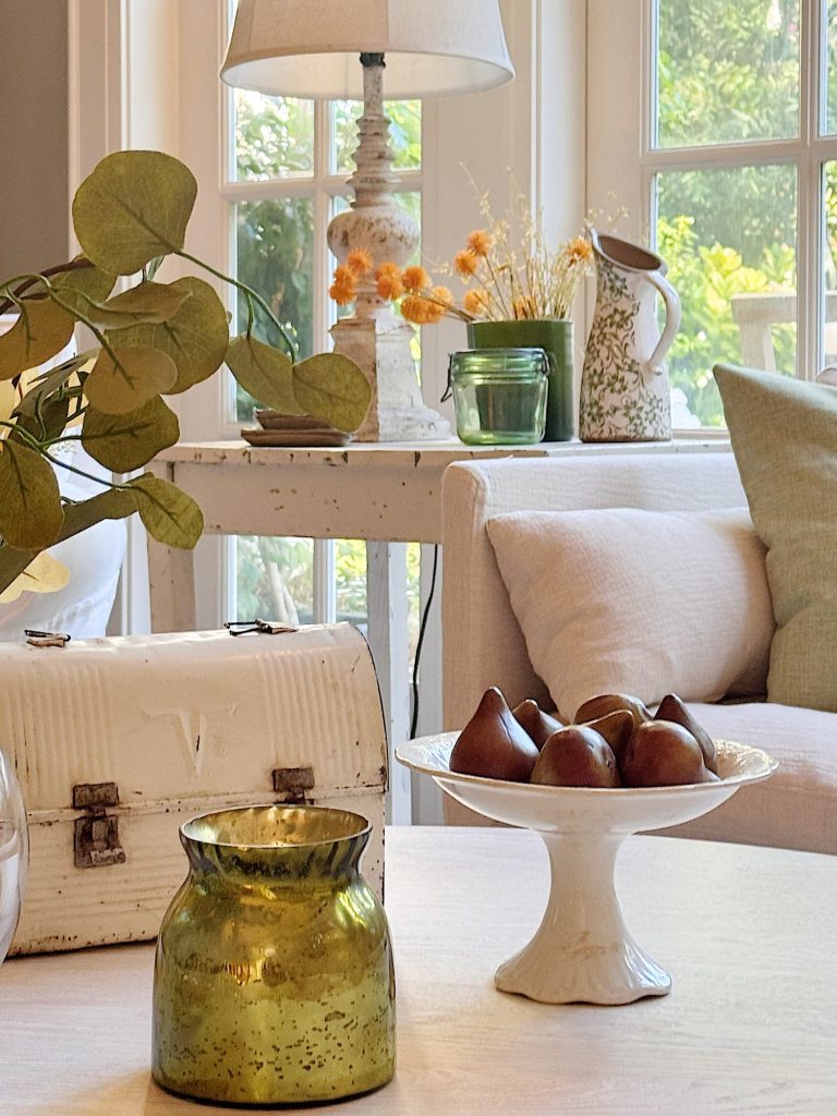 A living room scene with a white table holding a green glass vase, a white metal box, a pedestal plate with brown objects, and eucalyptus leaves, with a sofa and lamps in the background.