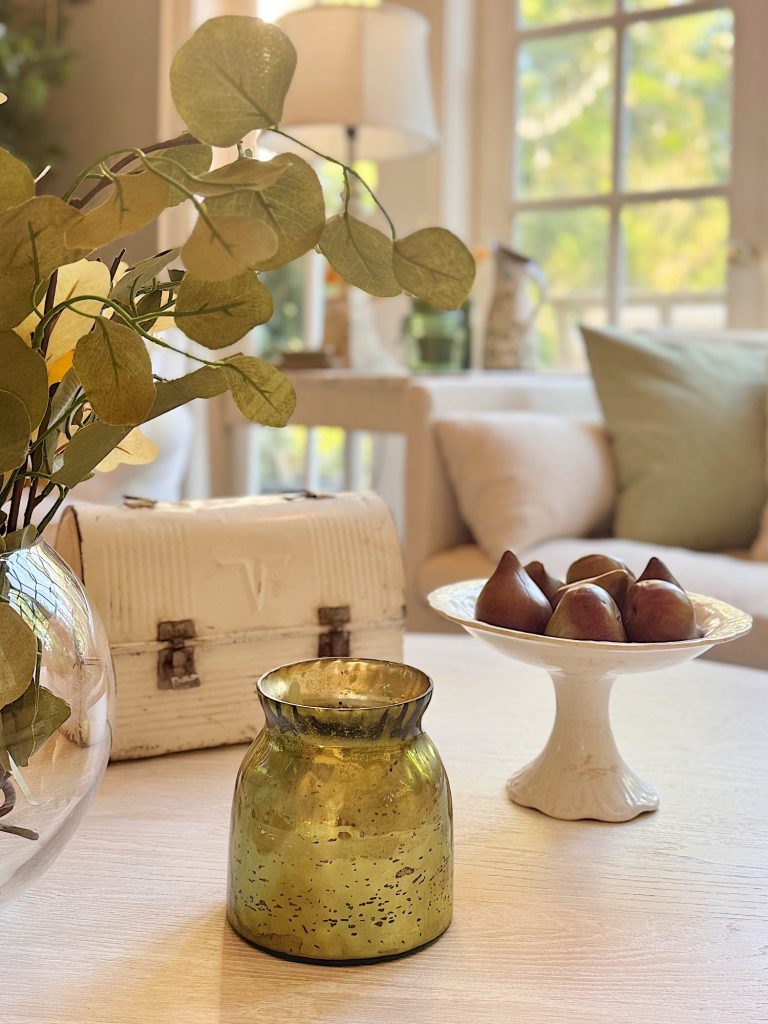 A gold-colored candle holder, a white pedestal dish with chocolates, and a vase with green leaves on a coffee table in a sunlit living room.