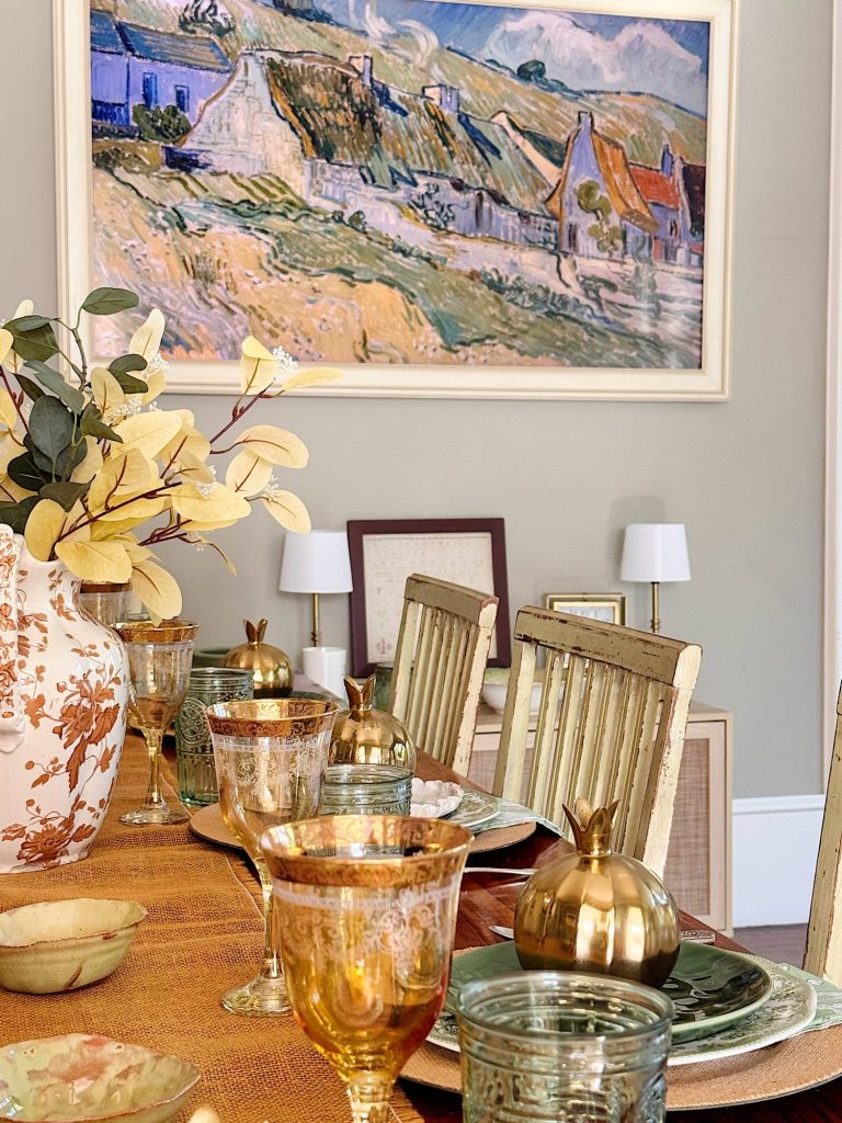 A dining table set with ornate glassware, gold accents, and a floral vase, with a large landscape painting and framed art on the wall in the background.