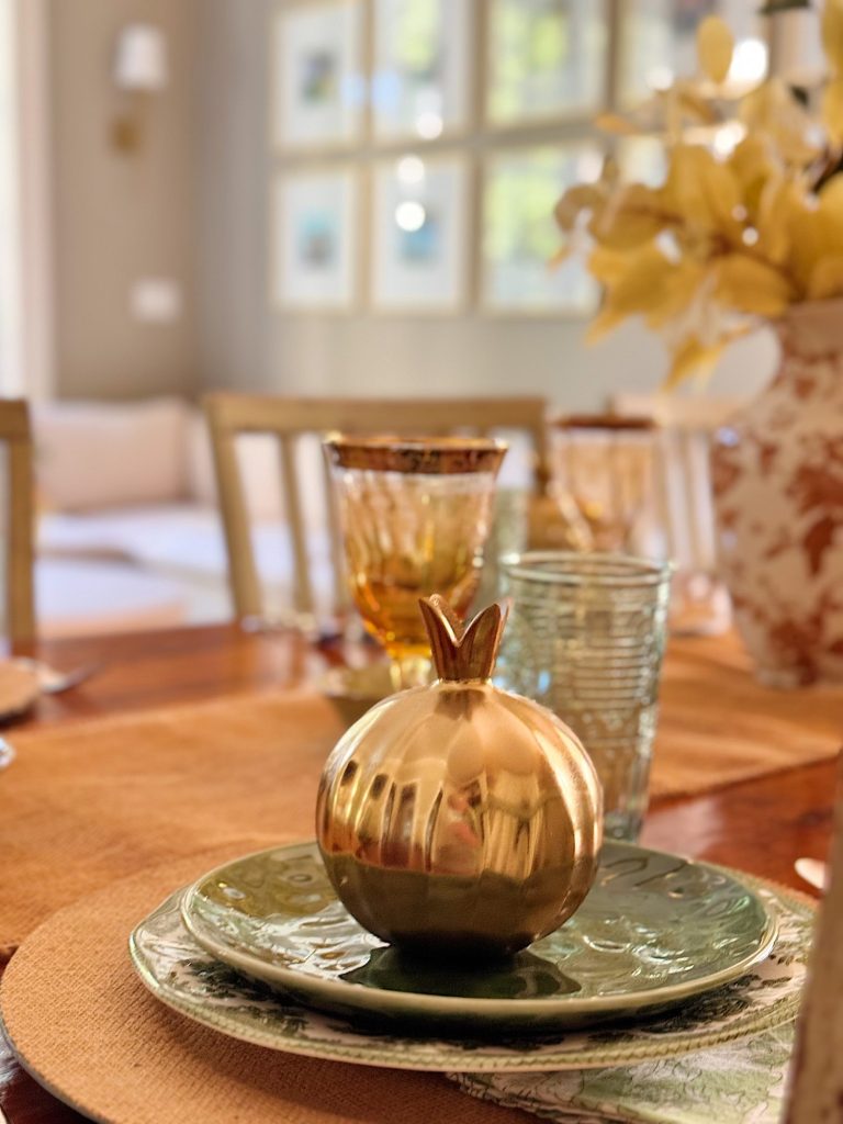 A dining table set with a decorative gold pomegranate on a green plate, surrounded by glassware and a floral vase in a sunlit room.