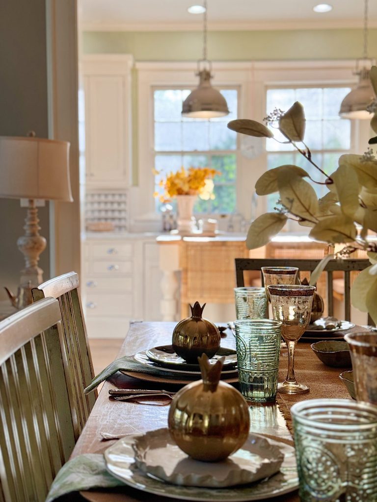A dining table is set with plates, green glassware, napkins, and gold pomegranate ornaments; a bright kitchen with flowers is visible in the background.