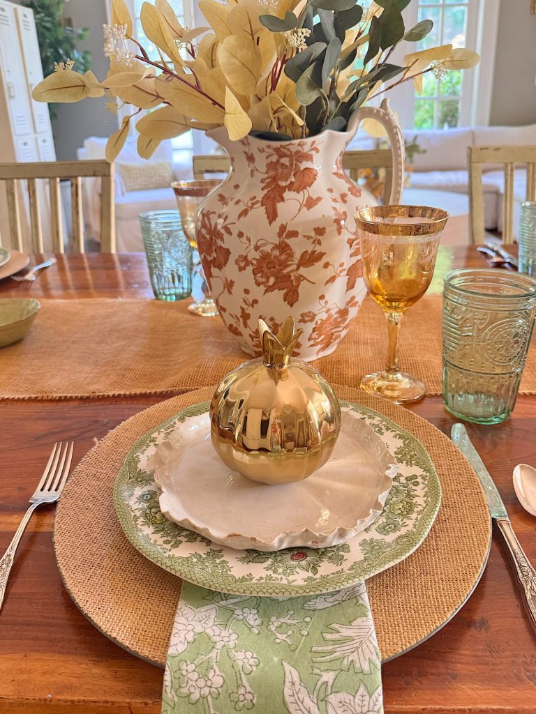 A table setting with a green and white floral plate, burlap placemat, green napkin, and a gold decorative apple, with a floral vase and glasses in the background.