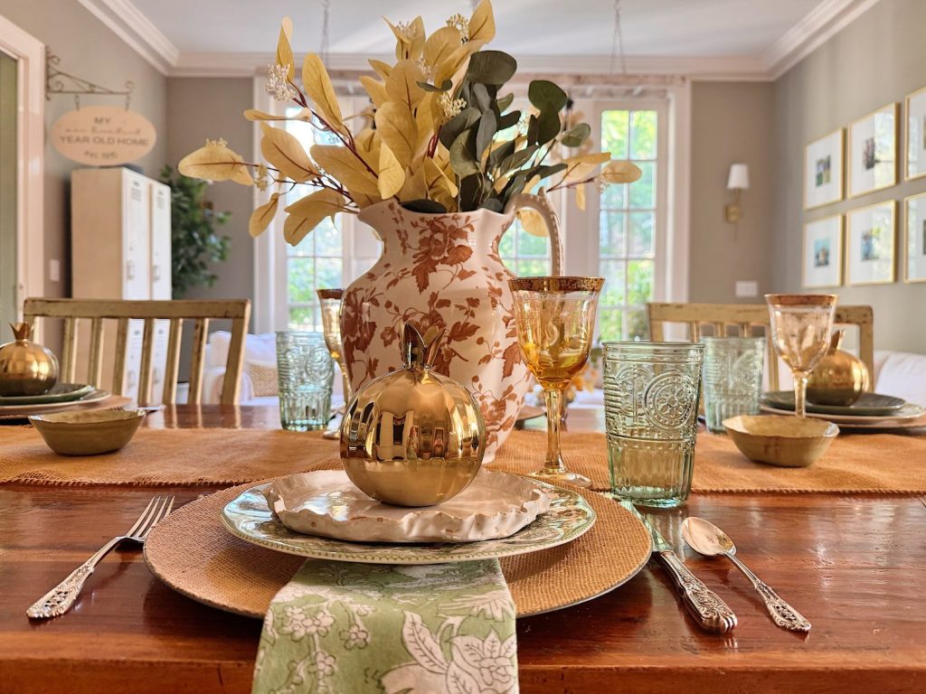 A dining table set with green and gold glassware, gold pumpkins on plates, a large floral vase centerpiece, and green napkins, in a sunlit room with beige walls and framed art.