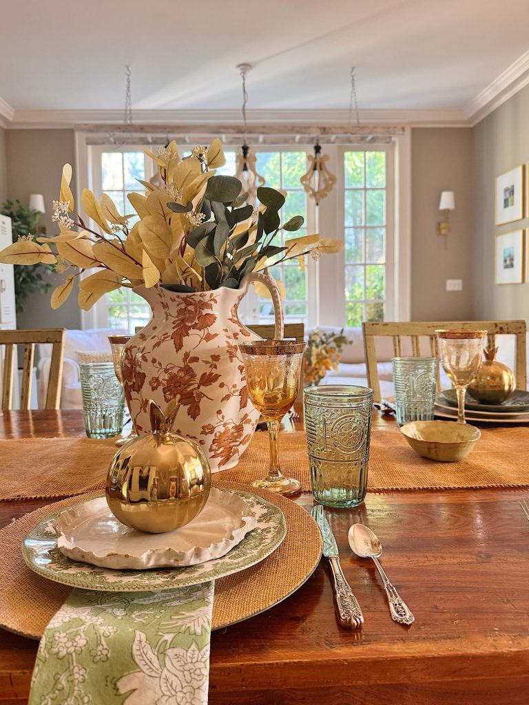 A dining table set with floral plates, gold cutlery, green glasses, gold goblets, a ceramic vase with foliage, and a gold apple ornament, in a sunlit room with large windows.