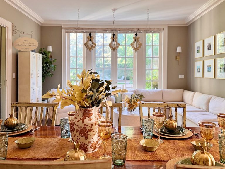A dining table set with dishes and glassware is in the foreground; a large window, white sofa, and wall art are visible in the bright living room beyond.