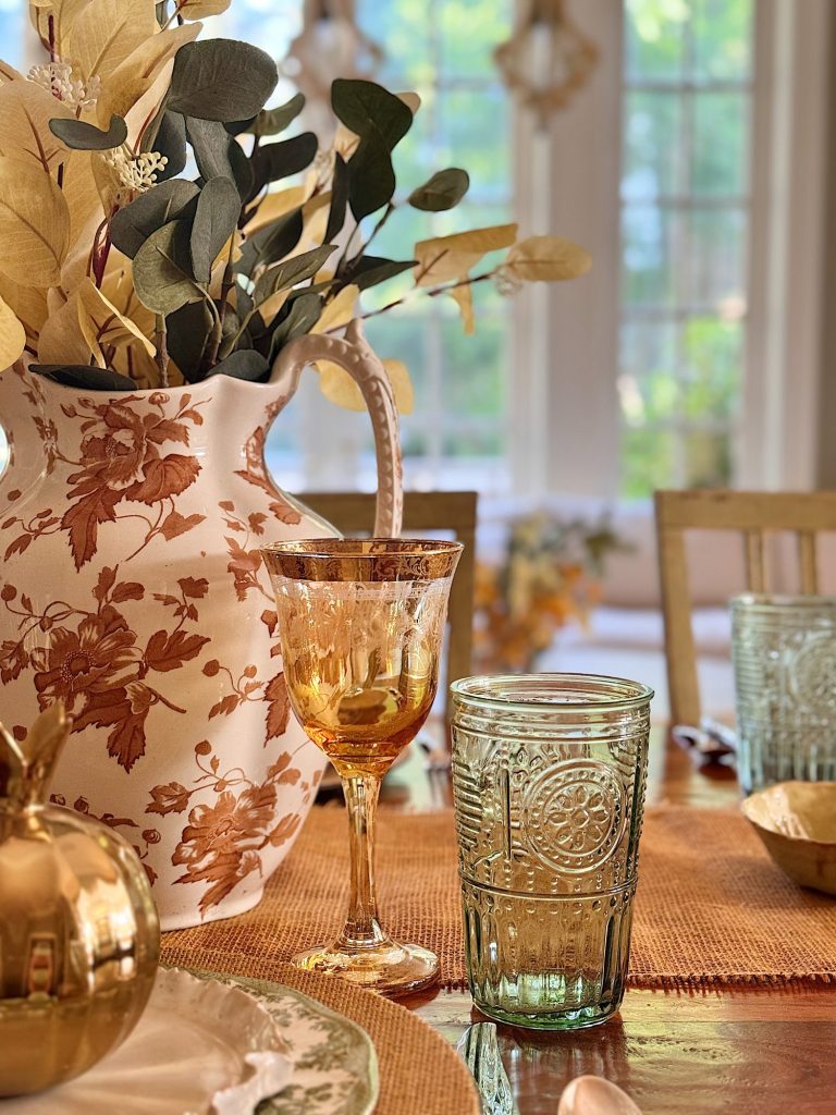 A dining table set with an amber goblet, patterned glass, floral pitcher with dried foliage, and gold decorative items, with sunlight streaming through large windows in the background.