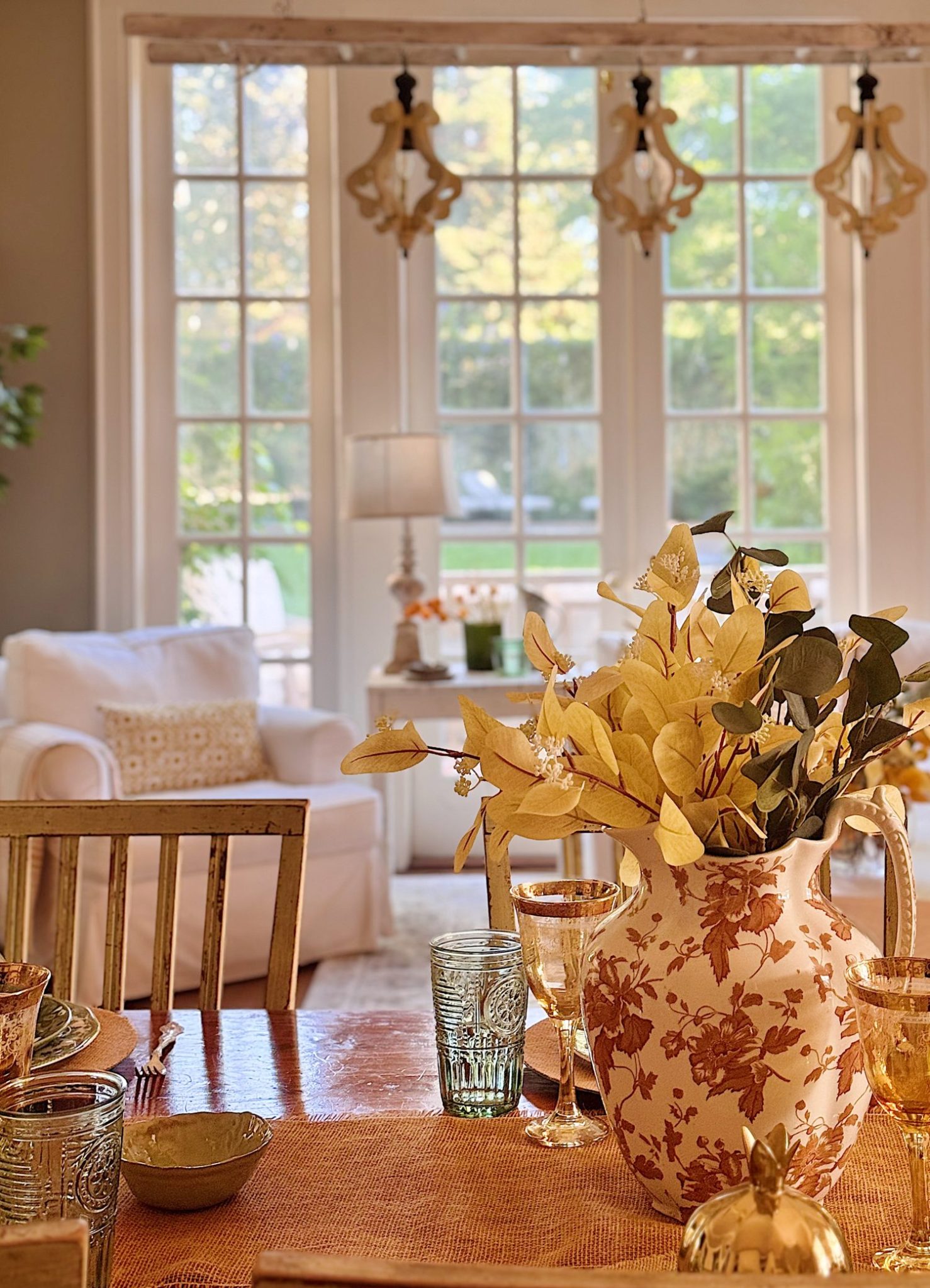 A floral pitcher with yellow leaves sits on a set dining table in a sunlit room with large windows, a white armchair, glassware, and a lamp in the background.