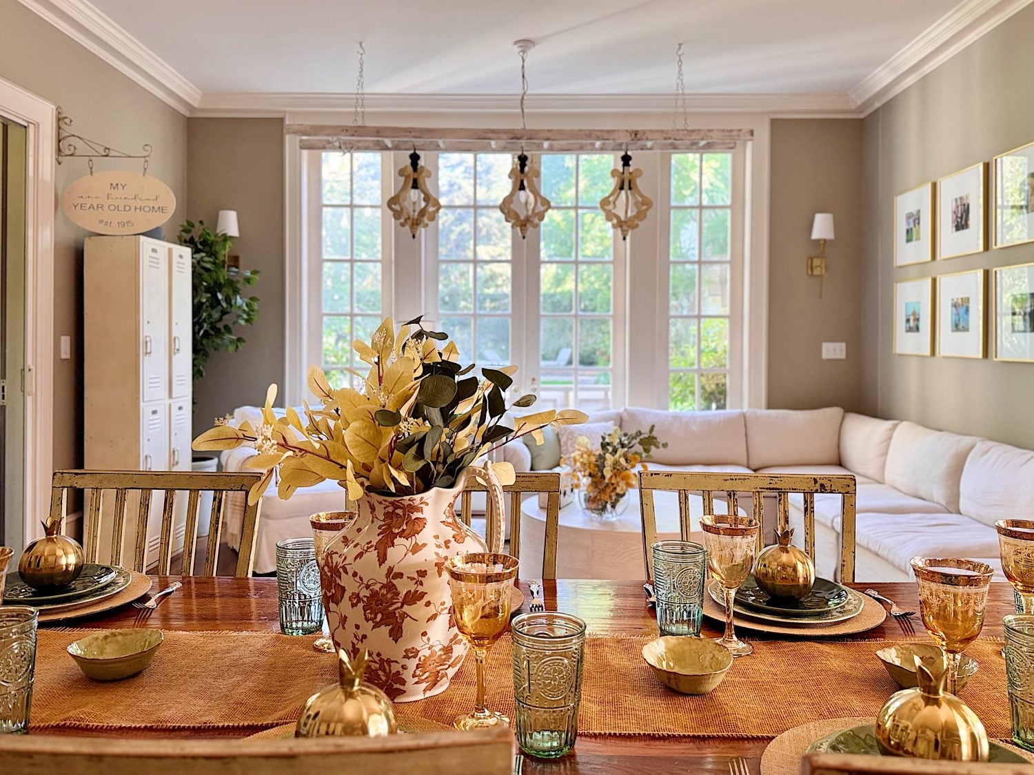 A dining table set with dishes and glassware is in the foreground; a large window, white sofa, and wall art are visible in the bright living room beyond.