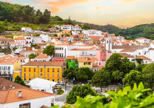A townscape with colorful buildings, trees, and hills in the background under a partly cloudy sky at sunset.