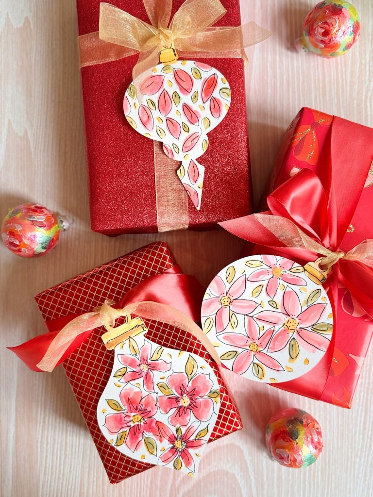 Two red gift boxes with pink ribbons and floral gift tags are displayed on a light wood surface, alongside two round floral-wrapped candies.