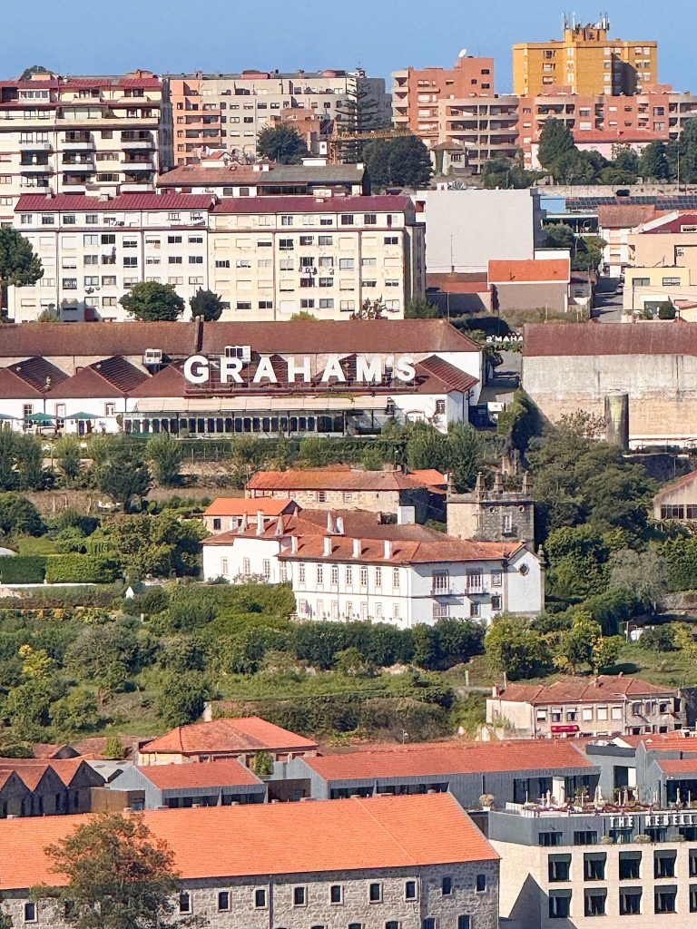 A large building with "GRAHAM'S" written on the roof sits on a hillside, surrounded by other buildings and greenery in an urban area.