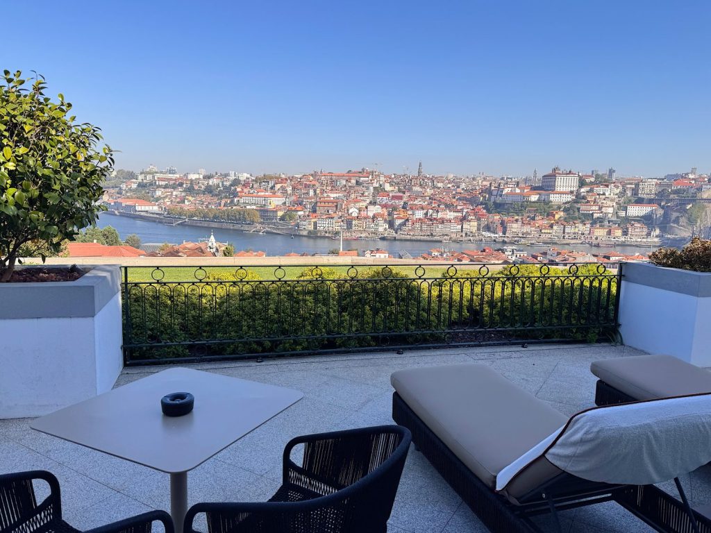 Terrace with lounge chairs and table overlooking a cityscape across a river under a clear blue sky.