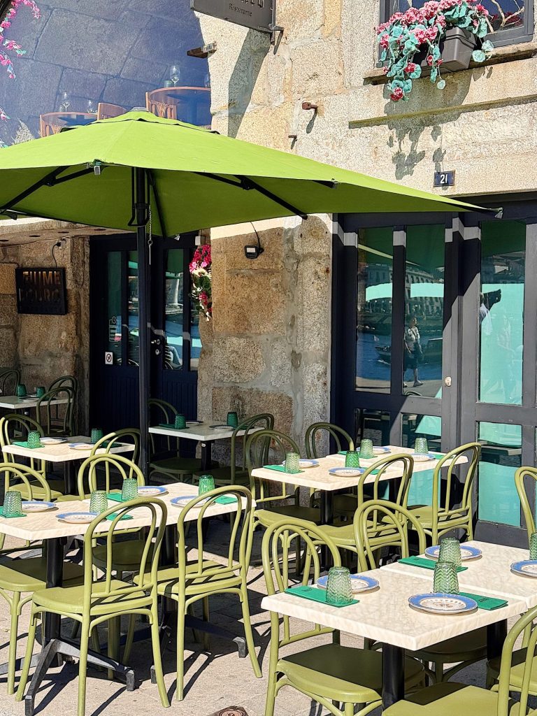 Outdoor café with green chairs and tables, each set with green glass cups and woven placemats, shaded by a large green umbrella beside a stone building with flower boxes.