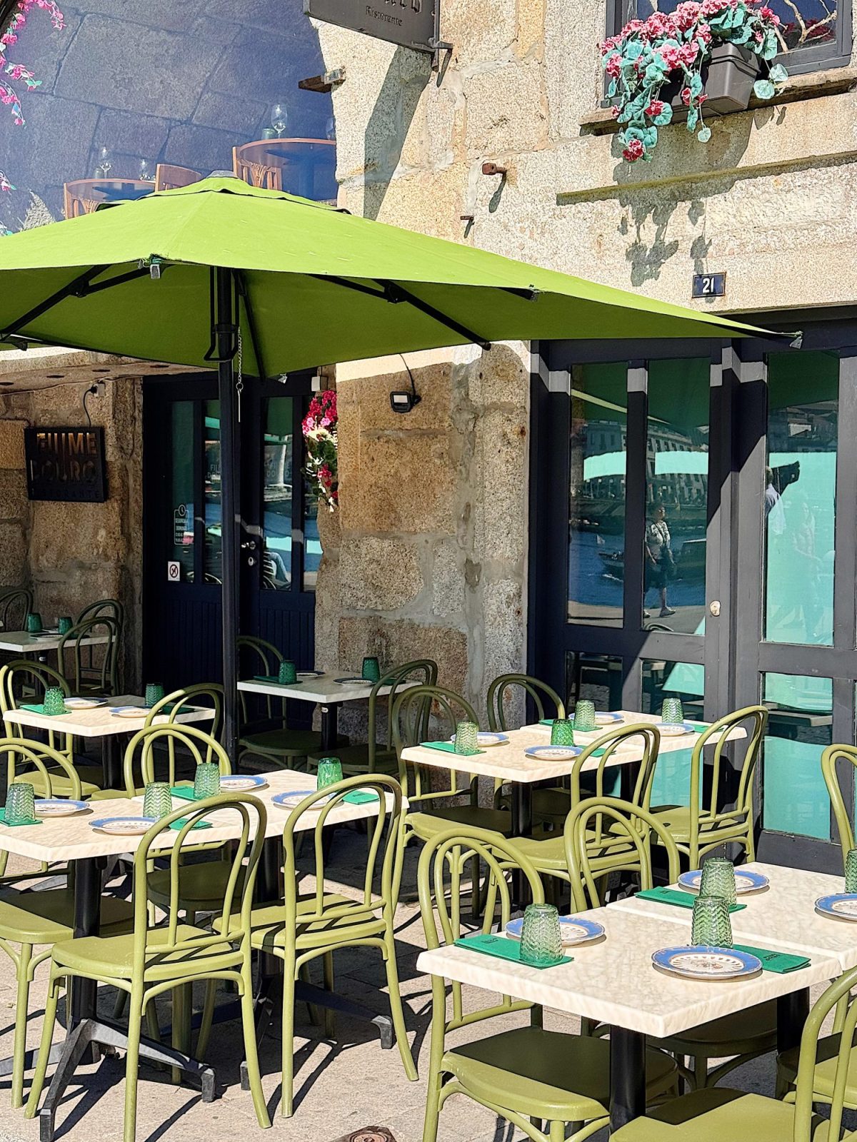 Outdoor caf&eacute; with green chairs and tables, each set with green glass cups and woven placemats, shaded by a large green umbrella beside a stone building with flower boxes.