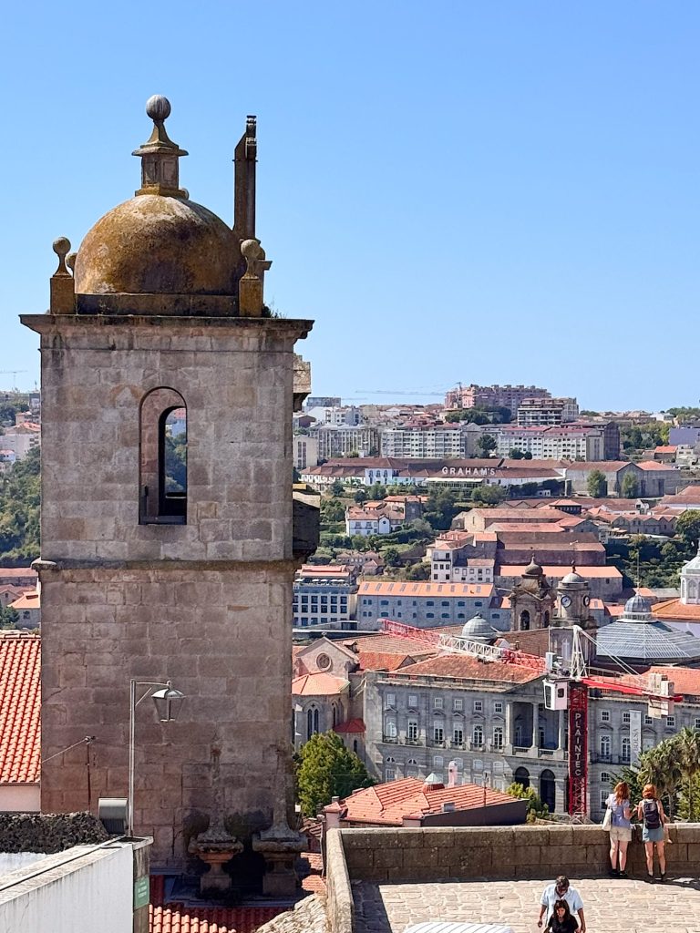 Stone bell tower with a dome overlooks a cityscape with red-roofed buildings, modern apartments, and people walking along a stone pathway under a clear blue sky.