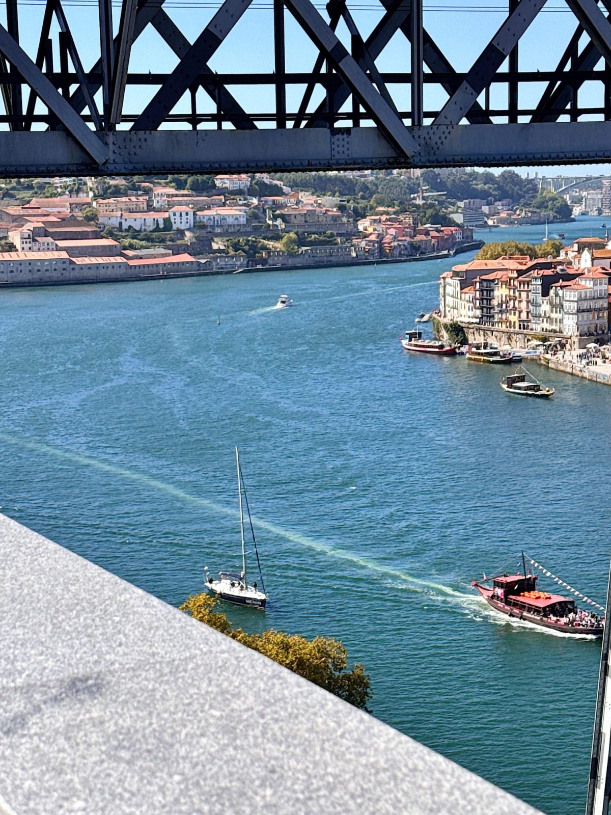 View of a river with boats and buildings along the shore, seen under a steel bridge structure on a sunny day.