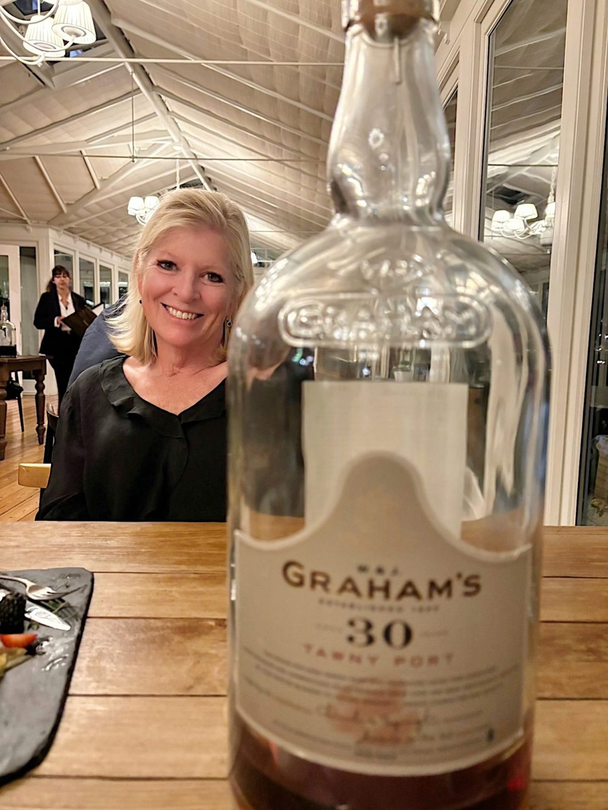 A woman sits at a table in a restaurant, smiling, with a large bottle of Graham&rsquo;s 30 Year Tawny Port in the foreground, partially obscuring her.