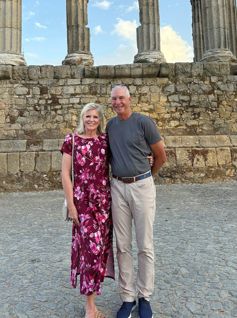 A woman in a floral dress and a man in a gray shirt and light pants stand together, smiling, in front of an ancient stone structure with tall columns.