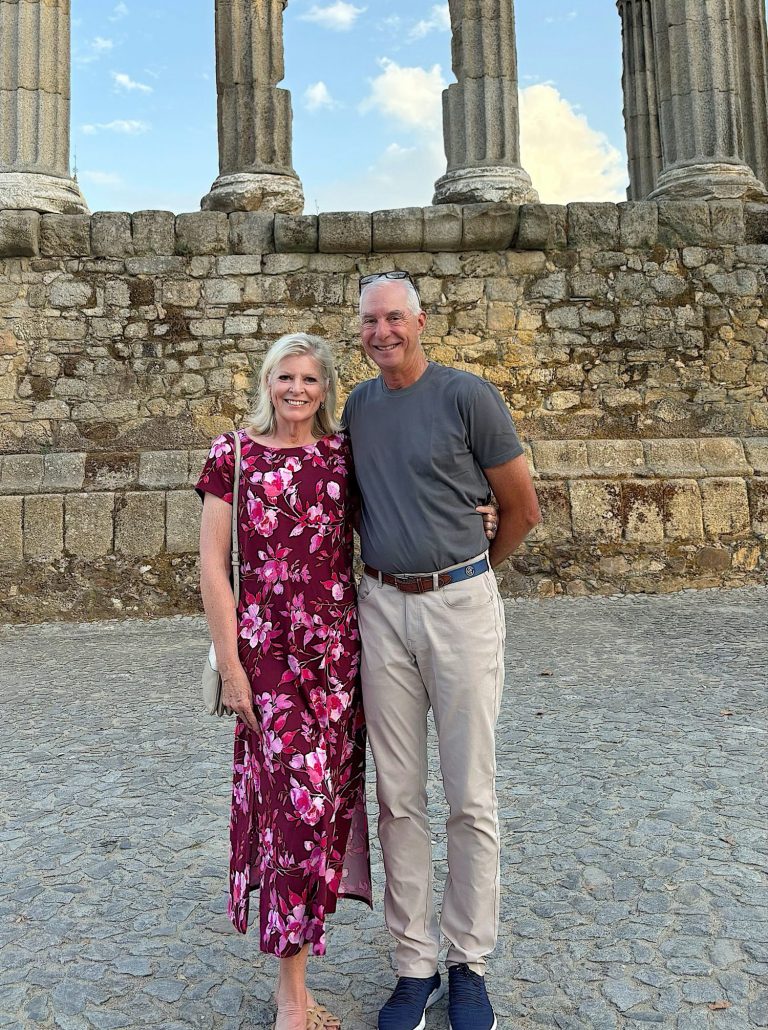 A woman in a floral dress and a man in a gray shirt and light pants stand together, smiling, in front of an ancient stone structure with tall columns.