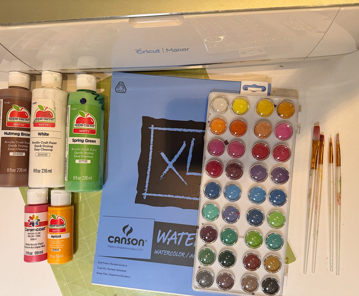 Art supplies including acrylic paint bottles, a watercolor paint set, paintbrushes, watercolor paper pad, and a Cricut Maker cutting machine are arranged on a table.