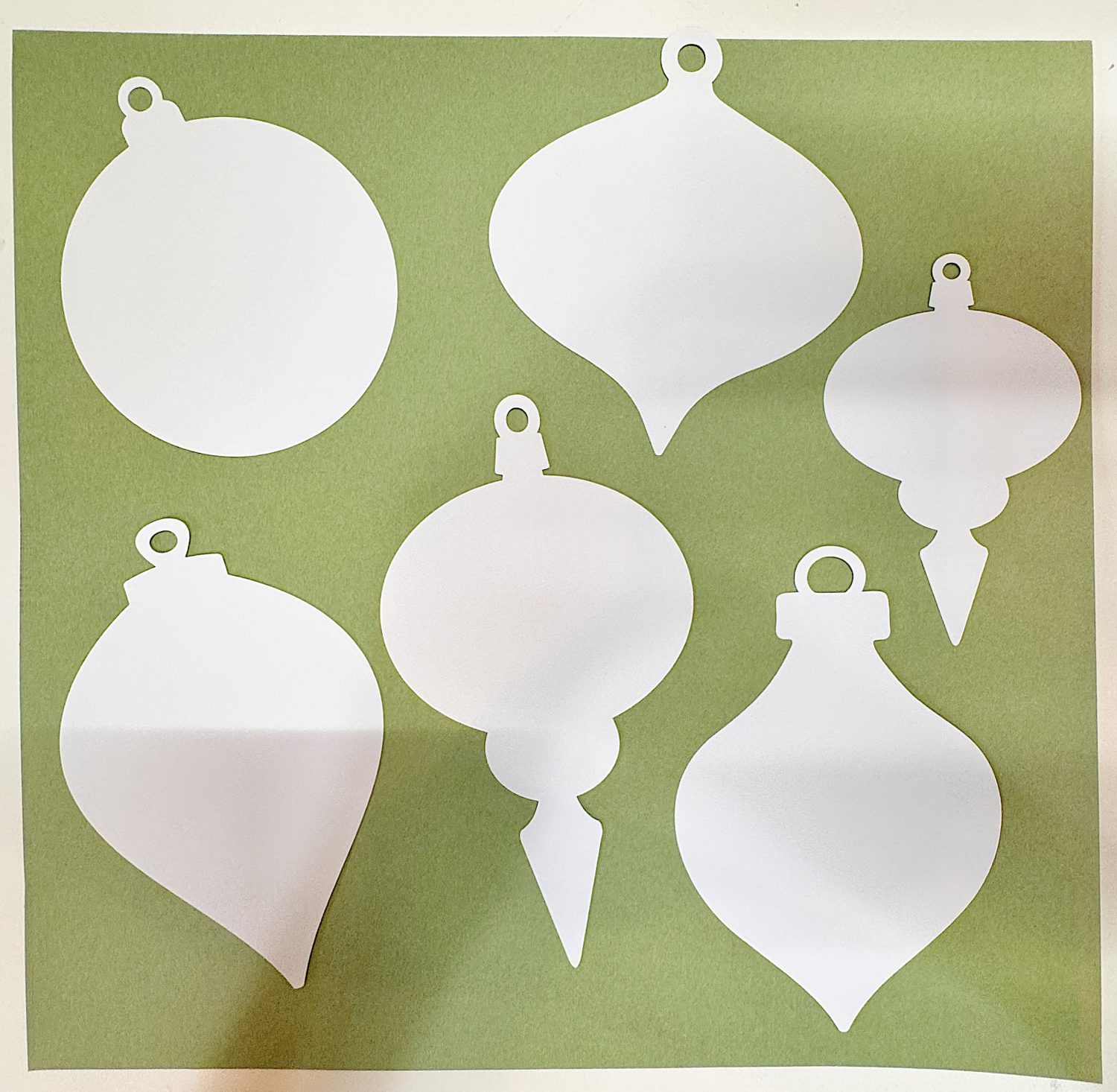 Six white paper cutouts in various Christmas ornament shapes are arranged on a green square background.