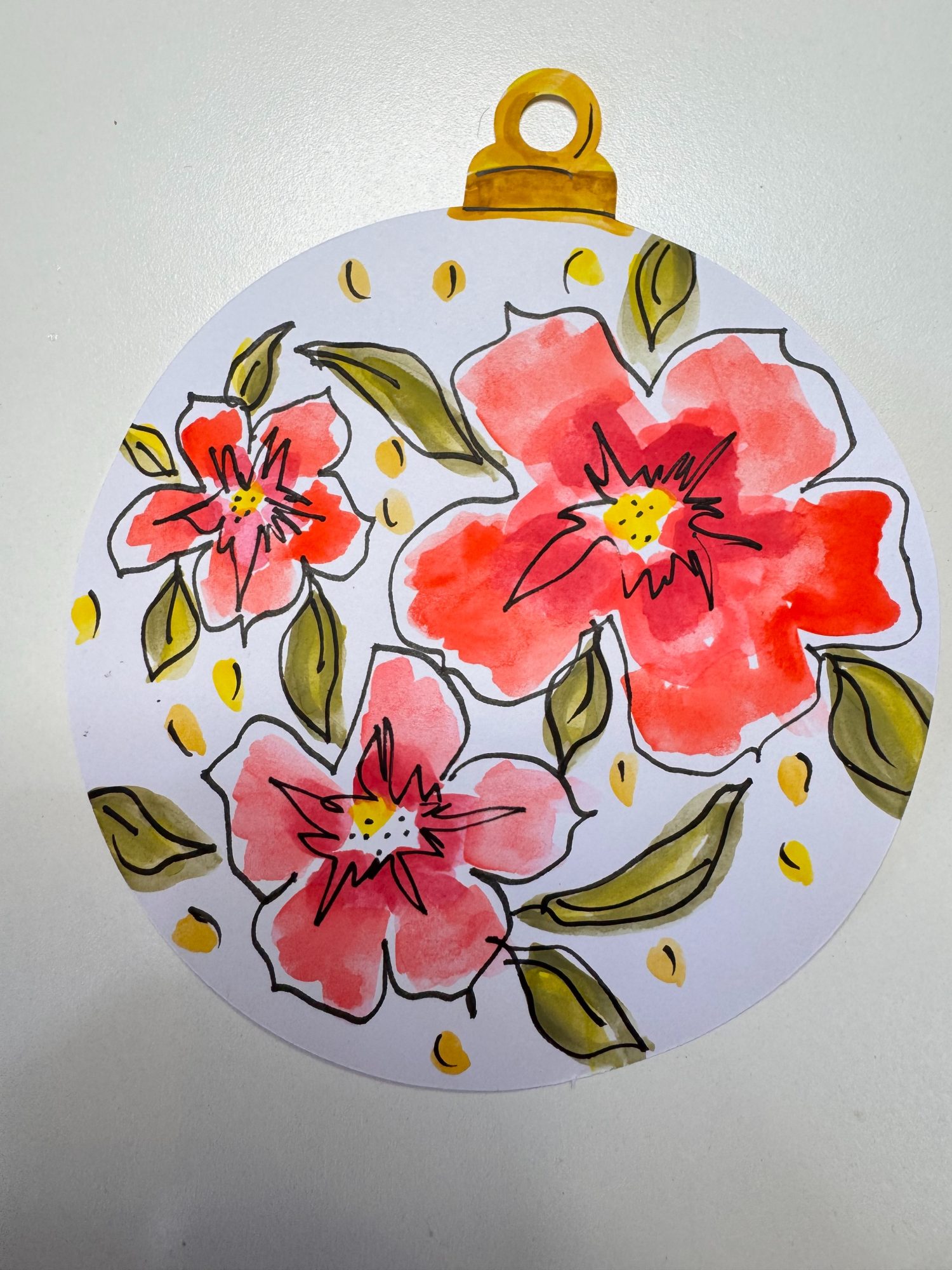 A round ornament with three red and pink flowers, green leaves, and yellow dots, drawn on white paper with a yellow loop at the top.