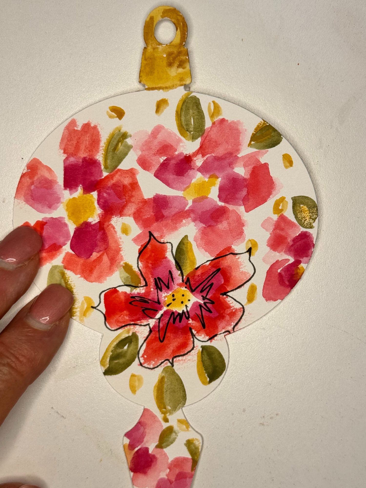 A hand holds a paper ornament painted with red and pink flowers, green leaves, and a yellow top, placed on a white surface.