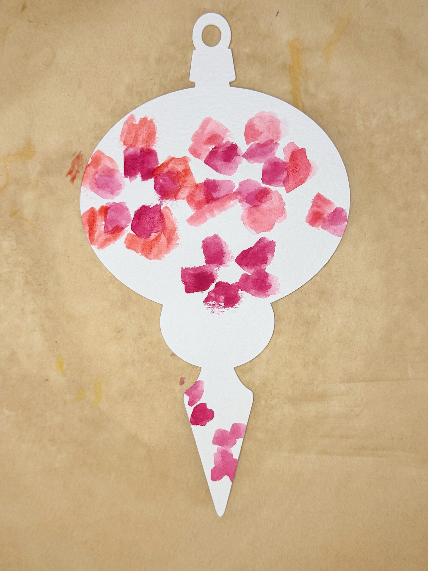 A white ornament-shaped cutout with red and pink watercolor blotches painted on it, placed on a light brown surface.